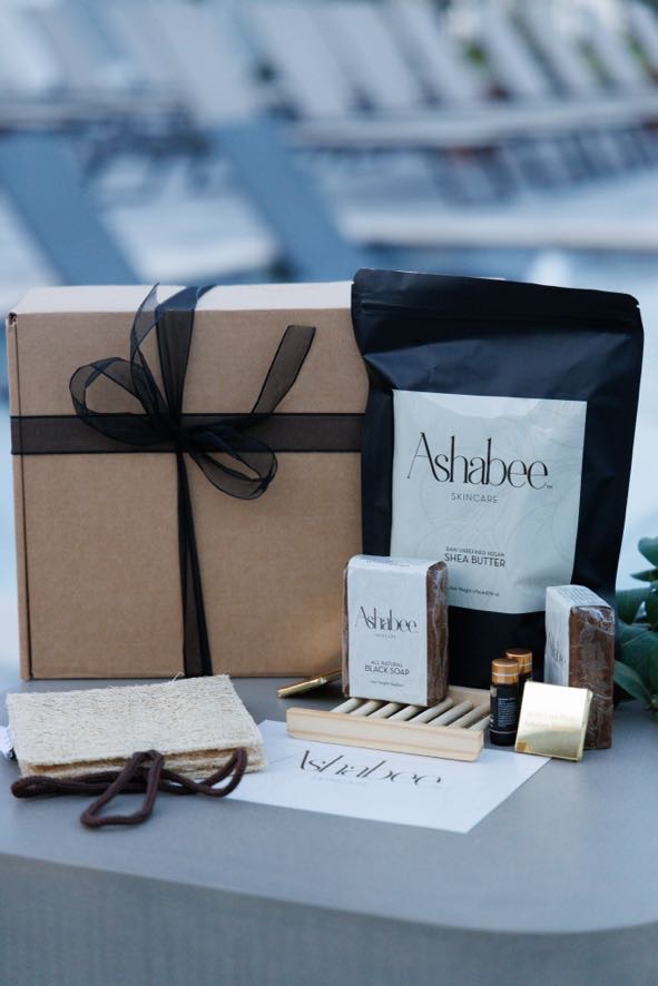 An Ashabee gift box tied with black ribbon, beside shea butter, black soap, botanical oil and a loofah